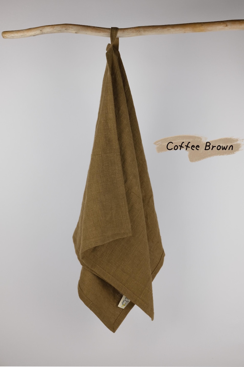 Puede incluir: Un pa&ntilde;o de cocina color marr&oacute;n caf&eacute; con una textura tejida visible que cuelga de una vara de madera. La tela tiene una peque&ntilde;a etiqueta cosida en la esquina inferior. El texto Coffee Brown est&aacute; impreso en una fuente cursiva oscura a la derecha.