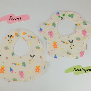 Pu&ograve; includere: Due bavaglini in tessuto color bianco sporco con un motivo colorato di piccoli orsi, conigli e forme. Uno &egrave; rotondo e l'altro &egrave; smerlato. Il testo "Round" appare in rosa e "Scalloped" in verde vicino ai rispettivi bavaglini.