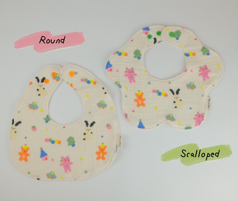 Pu&ograve; includere: Due bavaglini in tessuto color bianco sporco con un motivo colorato di piccoli orsi, conigli e forme. Uno &egrave; rotondo e l'altro &egrave; smerlato. Il testo "Round" appare in rosa e "Scalloped" in verde vicino ai rispettivi bavaglini.