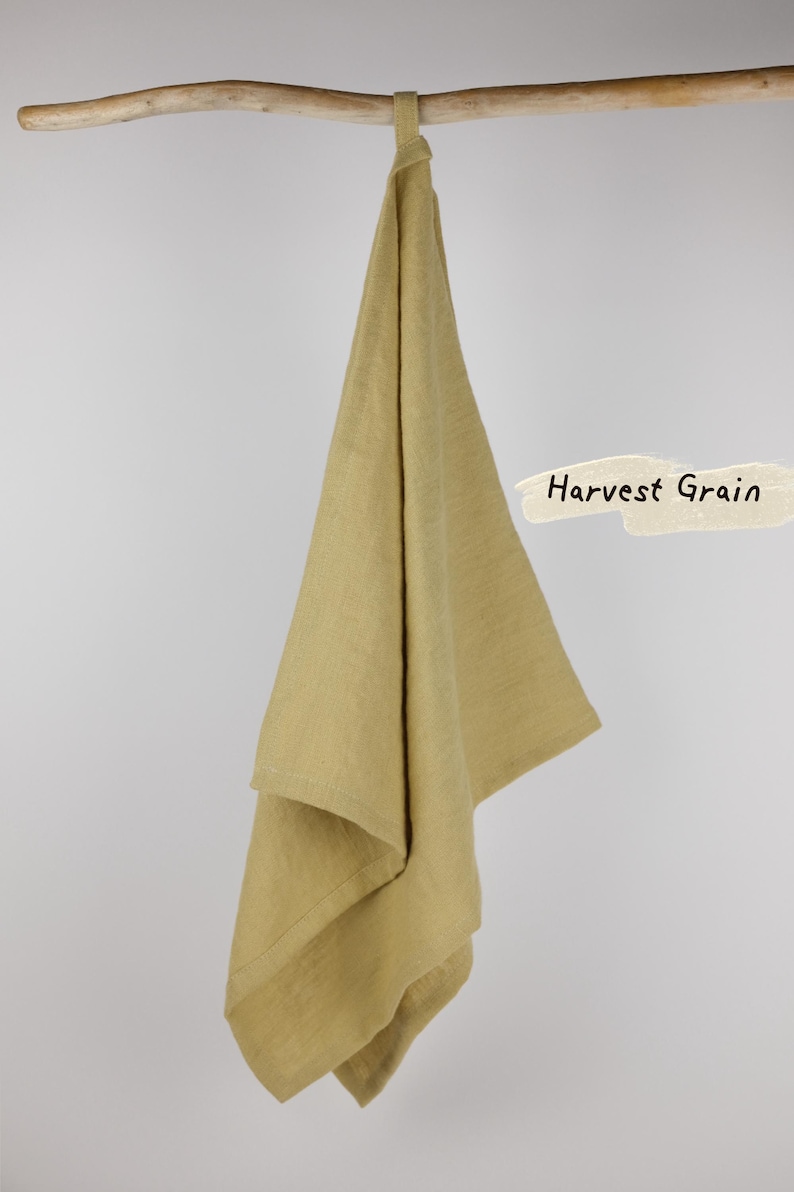 Puede incluir: Un pa&ntilde;o de cocina de color verde oliva claro liso con un peque&ntilde;o lazo para colgar en la parte superior. El pa&ntilde;o est&aacute; colgado sobre una rama de madera horizontal. A la derecha, hay un elemento gr&aacute;fico de color crema con el texto Harvest Grain.