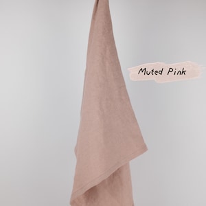 Puede incluir: Un pa&ntilde;o de cocina de color rosa apagado colgado de una rama de madera. La tela tiene una textura tejida visible y una peque&ntilde;a etiqueta rectangular en la esquina inferior. El texto Muted Pink est&aacute; escrito en negro sobre un fondo rosa claro.