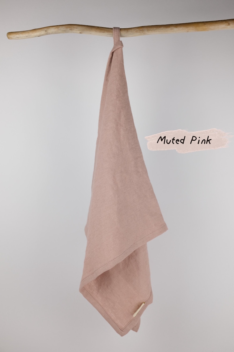 Puede incluir: Un pa&ntilde;o de cocina de color rosa apagado colgado de una rama de madera. La tela tiene una textura tejida visible y una peque&ntilde;a etiqueta rectangular en la esquina inferior. El texto Muted Pink est&aacute; escrito en negro sobre un fondo rosa claro.