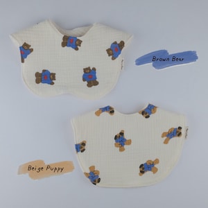 Pu&ograve; includere: Due bavaglini in tessuto color bianco sporco con superficie testurizzata. Uno presenta un motivo ripetuto di orsi bruni con camicie blu, etichettato Brown Bear. L'altro presenta un motivo ripetuto di cuccioli beige con camicie blu, etichettato Beige Puppy.