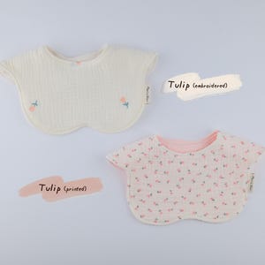 Pu&ograve; includere: Due bavaglini arrotondati per neonati in tessuto testurizzato. Uno &egrave; bianco sporco con piccoli tulipani rosa ricamati e steli verdi. L'altro &egrave; bianco con una fitta stampa di tulipani rosa. Entrambi hanno un'etichetta laterale Mari & Nui.