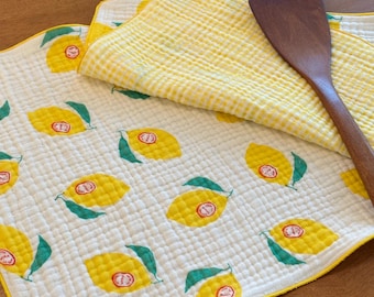 Può includere: Un panno in tessuto trapuntato bianco e giallo con un motivo ripetuto di limoni con foglie verdi e una piccola etichetta bianca con la scritta Mari & Nari, accompagnato da una spatola in legno marrone scuro sull'angolo piegato.