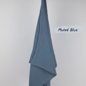 Puede incluir: Un pa&ntilde;o de cocina de color azul apagado colgado de una rama de madera. La tela tiene una textura tejida visible y un borde con dobladillo simple. Un gr&aacute;fico de pincelada azul claro junto al pa&ntilde;o muestra el texto negro escrito a mano Muted Blue.