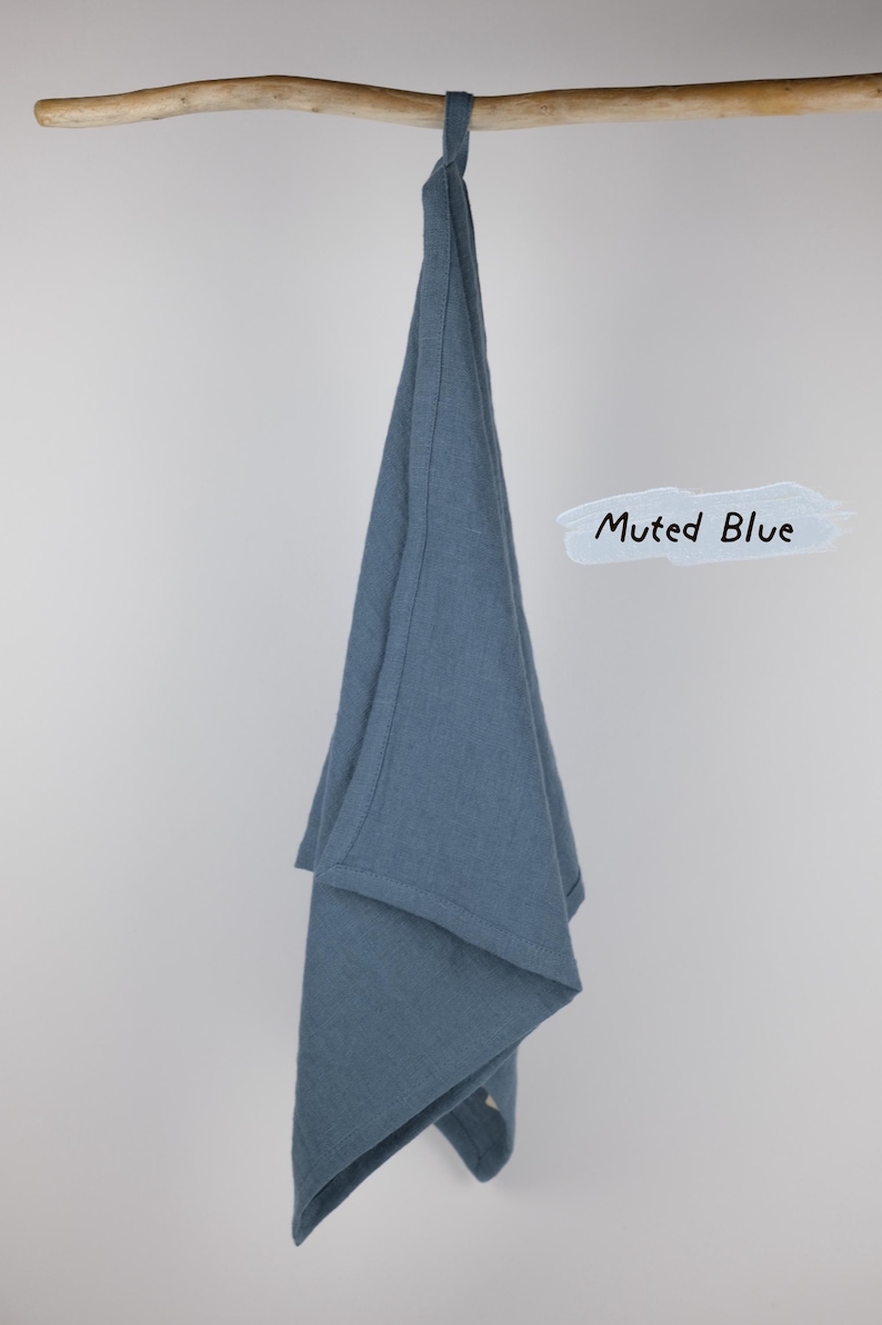 Puede incluir: Un pa&ntilde;o de cocina de color azul apagado colgado de una rama de madera. La tela tiene una textura tejida visible y un borde con dobladillo simple. Un gr&aacute;fico de pincelada azul claro junto al pa&ntilde;o muestra el texto negro escrito a mano Muted Blue.