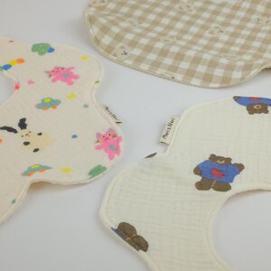 Pu&ograve; includere: Tre bavaglini in tessuto con bordi curvi. Uno presenta un motivo a quadretti beige e bianco con piccoli volti di orso. Un altro &egrave; color crema con gatti, stelle e auto colorate. Il terzo &egrave; color crema con orsi marroni che indossano maglioni blu, etichettato Mari & Nari.
