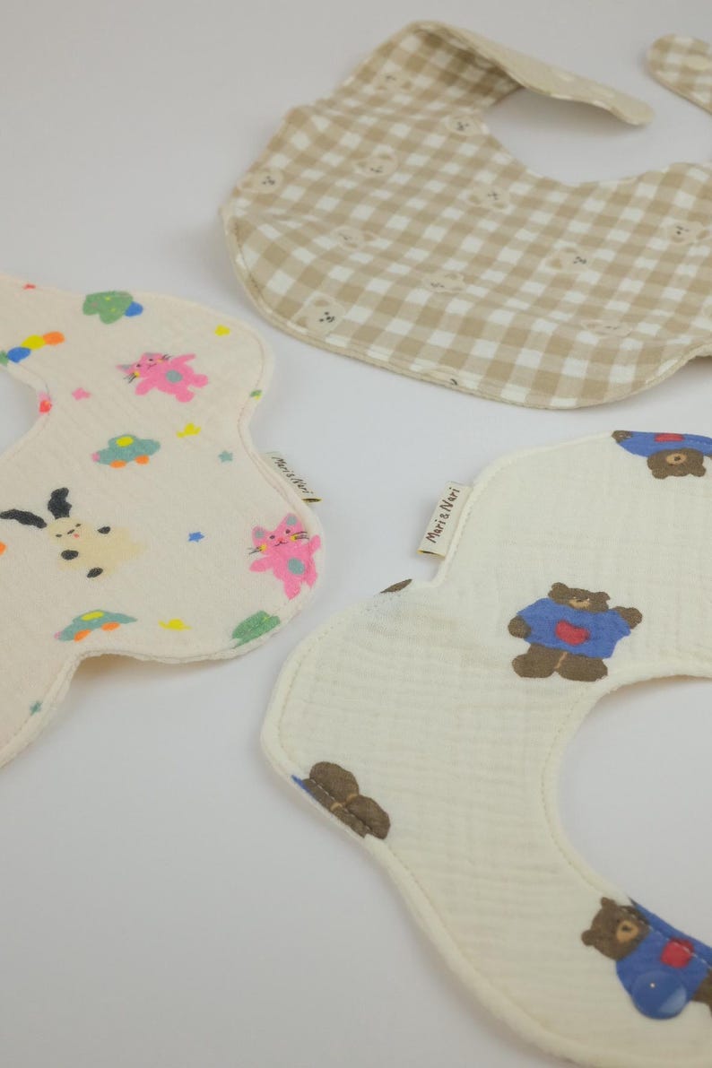 Pu&ograve; includere: Tre bavaglini in tessuto con bordi curvi. Uno presenta un motivo a quadretti beige e bianco con piccoli volti di orso. Un altro &egrave; color crema con gatti, stelle e auto colorate. Il terzo &egrave; color crema con orsi marroni che indossano maglioni blu, etichettato Mari & Nari.