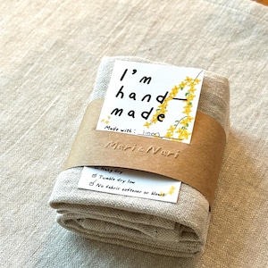 May include: A folded linen dish towel with a brown tag that reads "I'm hand made, Made with: linen, Marie Vari" and a white tag with care instructions: "Hang dry, Tumble dry low, No fabric softener or bleach".