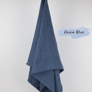 Puede incluir: Un pa&ntilde;o de cocina color azul denim con un lazo para colgar, drapeado sobre una rama de madera. La tela tiene una textura tejida visible y un color azul profundo s&oacute;lido. A la derecha, un gr&aacute;fico de trazo azul claro contiene el texto Denim Blue.