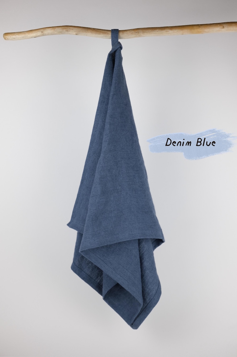Puede incluir: Un pa&ntilde;o de cocina color azul denim con un lazo para colgar, drapeado sobre una rama de madera. La tela tiene una textura tejida visible y un color azul profundo s&oacute;lido. A la derecha, un gr&aacute;fico de trazo azul claro contiene el texto Denim Blue.