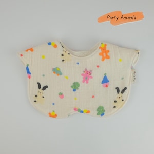 Pu&ograve; includere: Una bavaglino in tessuto color crema, di forma arrotondata, con un motivo colorato di piccoli animali, alberi e stelle. Il testo "Party Animals" appare in una grafica a pennellata arancione sopra il bavaglino, con un'etichetta "Maris Noel".