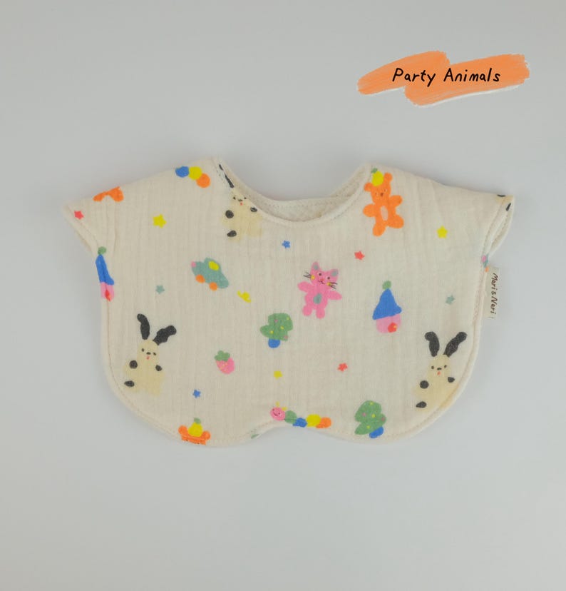 Pu&ograve; includere: Una bavaglino in tessuto color crema, di forma arrotondata, con un motivo colorato di piccoli animali, alberi e stelle. Il testo "Party Animals" appare in una grafica a pennellata arancione sopra il bavaglino, con un'etichetta "Maris Noel".