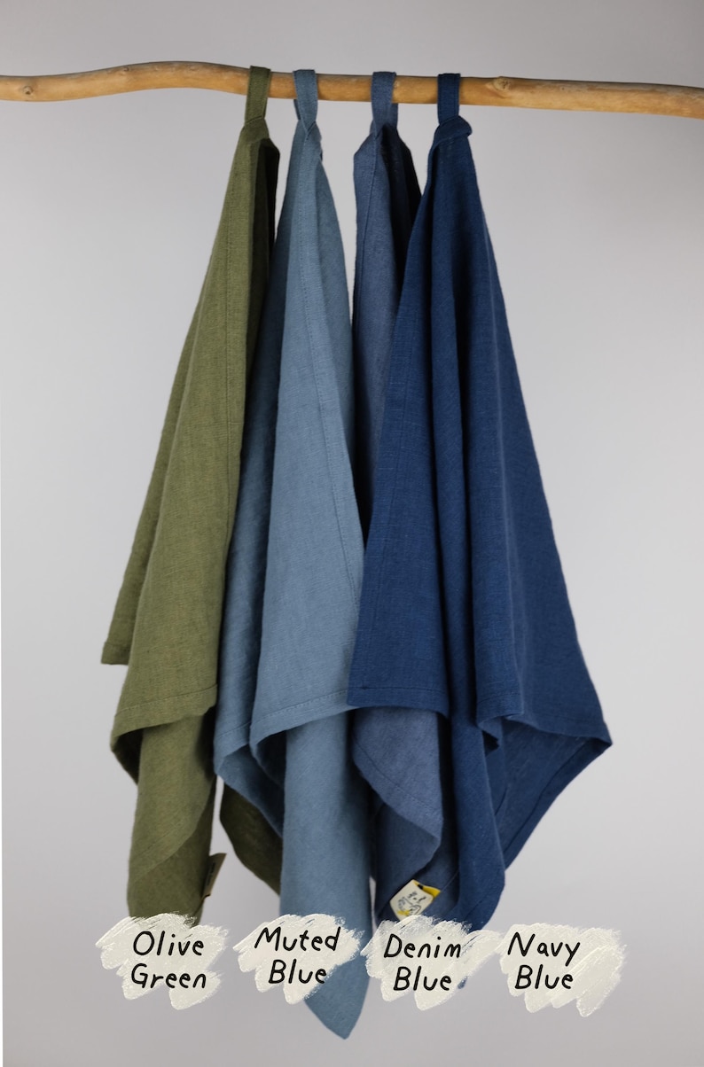 Puede incluir: Cuatro pa&ntilde;os de cocina rectangulares colgados de una barra de madera, cada uno con un lazo de tela en la parte superior. Los colores de izquierda a derecha est&aacute;n etiquetados como Olive Green, Muted Blue, Denim Blue y Navy Blue, con una etiqueta.