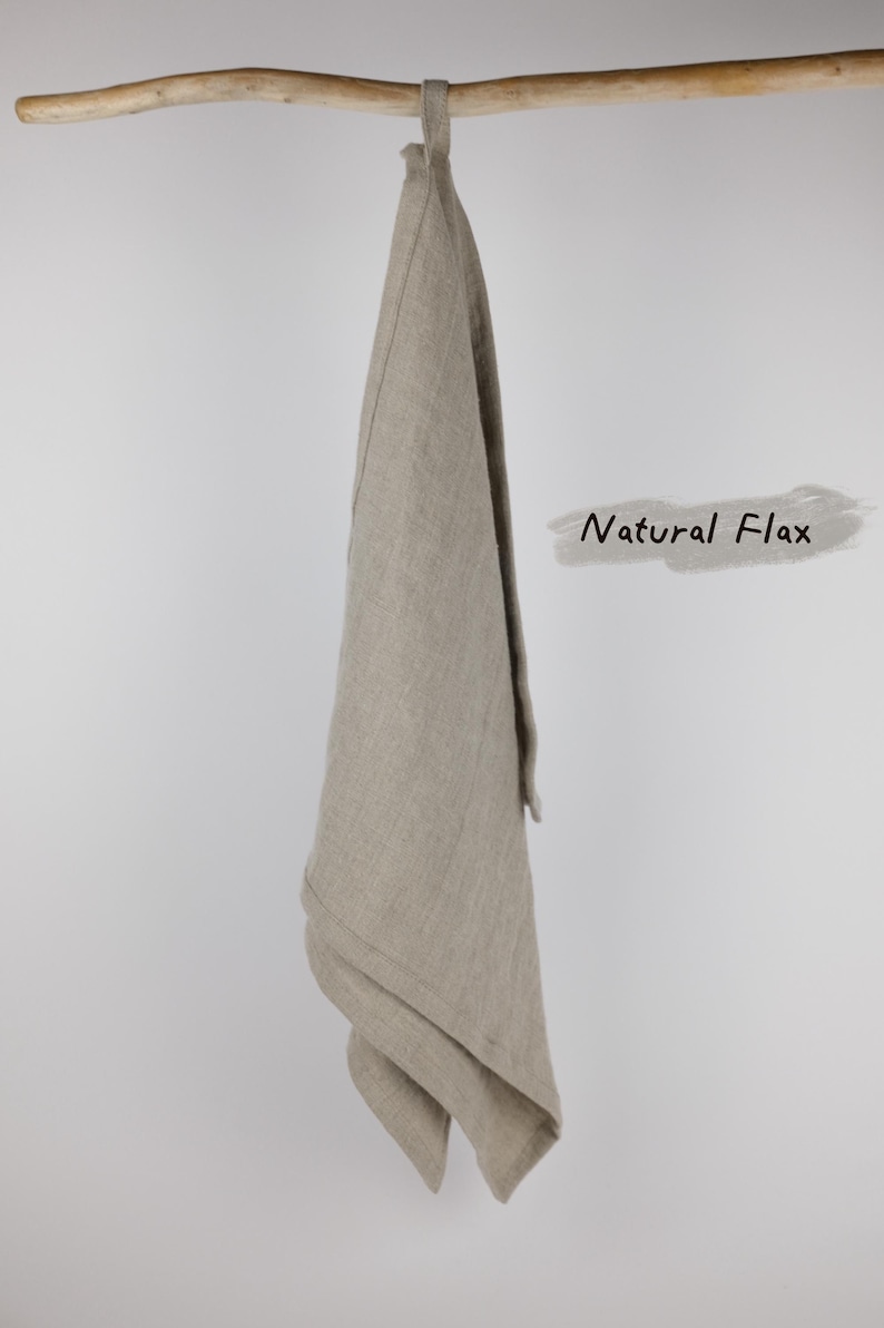 Puede incluir: Un pa&ntilde;o de cocina rectangular hecho de tela de lino de color beige claro que cuelga de una rama de madera. El pa&ntilde;o cuenta con un peque&ntilde;o lazo de tela en la parte superior para colgarlo. El texto Natural Flax est&aacute; impreso en negro a la derecha.