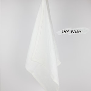 Puede incluir: Un pa&ntilde;o de cocina de color blanco roto colgado de una rama de madera. El pa&ntilde;o presenta un borde sencillo con dobladillo y una trabilla de tela en la parte superior para colgarlo. El texto "Off White" est&aacute; impreso en negro a la derecha.