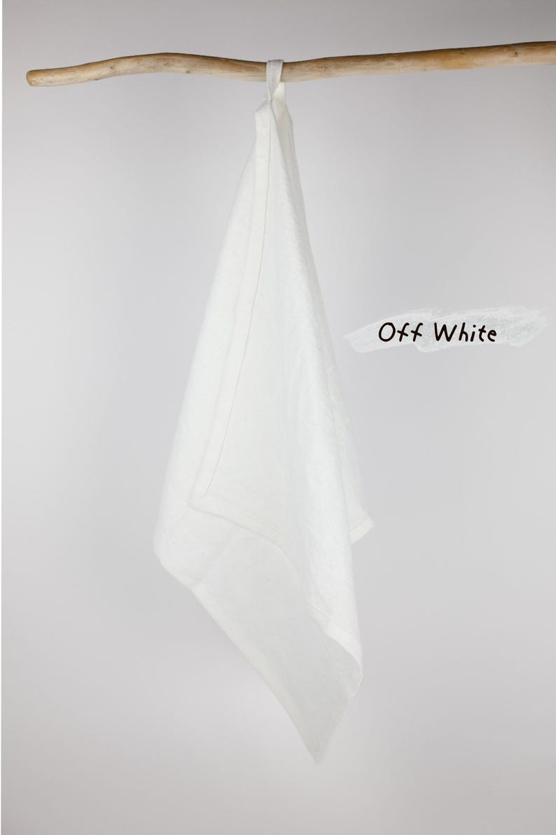 Puede incluir: Un pa&ntilde;o de cocina de color blanco roto colgado de una rama de madera. El pa&ntilde;o presenta un borde sencillo con dobladillo y una trabilla de tela en la parte superior para colgarlo. El texto "Off White" est&aacute; impreso en negro a la derecha.
