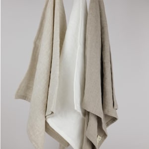 Puede incluir: Tres toallas de tela rectangulares colgadas de una varilla de madera. Las toallas est&aacute;n etiquetadas como White Dove, Off White y Natural Flax. Cada toalla presenta un lazo simple en la parte superior para colgar y un tono neutro.
