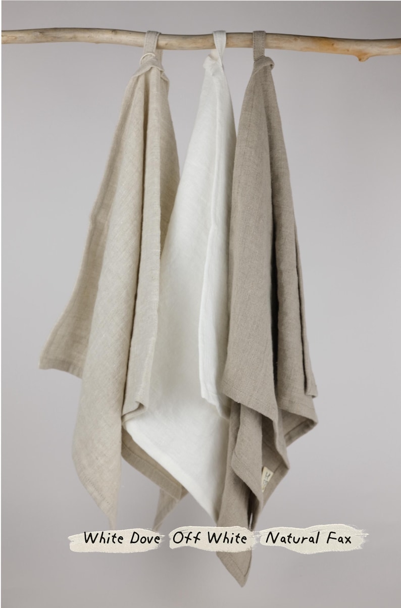 Puede incluir: Tres toallas de tela rectangulares colgadas de una varilla de madera. Las toallas est&aacute;n etiquetadas como White Dove, Off White y Natural Flax. Cada toalla presenta un lazo simple en la parte superior para colgar y un tono neutro.