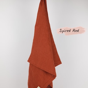 Puede incluir: Un pa&ntilde;o de cocina de color rojo especiado colgado de una varilla de madera. El tejido rectangular presenta una textura visible y una peque&ntilde;a etiqueta cuadrada en la esquina inferior. El texto Spiced Red aparece en negro sobre un fondo rosa.