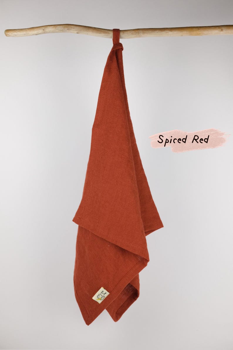 Puede incluir: Un pa&ntilde;o de cocina de color rojo especiado colgado de una varilla de madera. El tejido rectangular presenta una textura visible y una peque&ntilde;a etiqueta cuadrada en la esquina inferior. El texto Spiced Red aparece en negro sobre un fondo rosa.