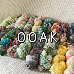 May include: A collection of numerous skeins of yarn in a wide variety of colours, including shades of green, pink, yellow, blue, and grey, with some featuring variegated patterns. The text OOAK is overlaid in white on the centre of the yarn pile.
