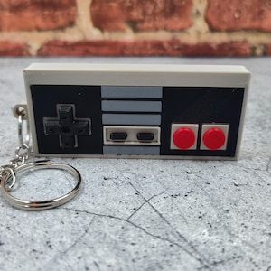 Puede incluir: Un llavero de color gris y negro con forma de mando de Nintendo, con botones rojos. El llavero tiene un anillo de metal plateado.
