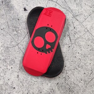 May include: A red and black fingerboard with a skull design. The fingerboard is red with a black skull graphic and black grip tape. The fingerboard is approximately 4 inches long.