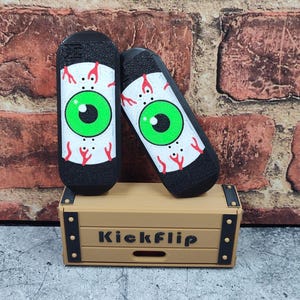 May include: Two black finger skateboards with an eye design, featuring a green iris and red veins, rest on a small wooden box. The box is tan with the word "Kickflip" printed on the front.