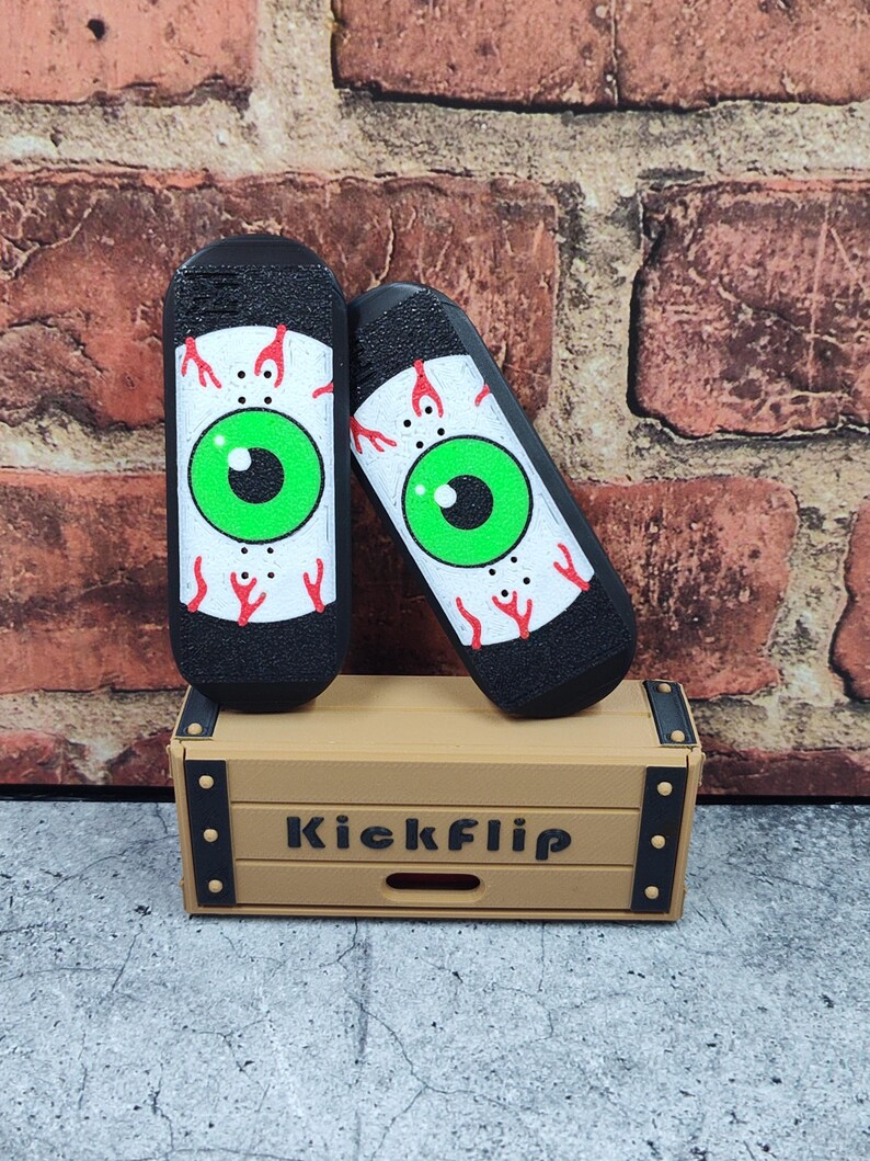 May include: Two black finger skateboards with an eye design, featuring a green iris and red veins, rest on a small wooden box. The box is tan with the word "Kickflip" printed on the front.