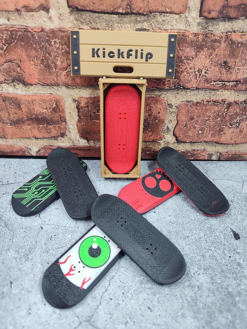 May include: A collection of miniature skateboards in various designs. One red board is displayed in a wooden box, and the word "Kickflip" is above it. Other boards feature designs like a circuit board, an eye, and a skull.