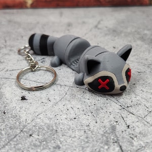 May include: A gray raccoon keychain with red X's for eyes. The keychain has a silver metal ring.