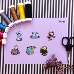 Könnte beinhalten: Sechs gestickte Aufnäher mit Horrorfilmfiguren, darunter Gestalten in blauen und schwarzen Outfits, angeordnet auf einer hellvioletten Fläche. Garnrollen in verschiedenen Farben, eine schwarze Schere und getrocknete violette Blumen liegen um die Aufnäher.