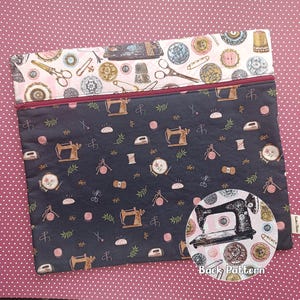 May include: A rectangular fabric pouch featuring a dark grey main section with a repeating pattern of sewing machines, irons, and yarn, and a light pink top band with sewing notions. A maroon zip secures the top, and a circular inset shows the back.