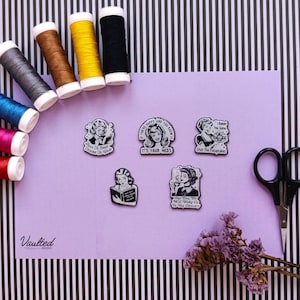 May include: Five black and white enamel pins featuring retro-style illustrations and text, including phrases like Born To Read Forced To Work, It's Your Meds, I Baked You Some Shut The Fucupcakes, and One Day We Will Wake Up To His Obituary.