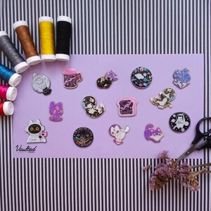 Puede incluir: Una colección de quince pequeños pines decorativos con gatos espaciales, planetas y tostadas, dispuestos sobre una superficie de color violeta claro. El texto Vaulted Stitches está impreso en la esquina inferior izquierda.