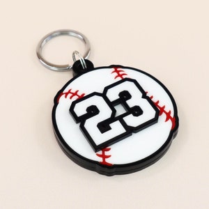 Könnte beinhalten: Ein weißer Baseball-Schlüsselanhänger mit schwarzen Nähten und der Nummer 23 in schwarzen Buchstaben. Der Schlüsselanhänger ist an einem silbernen Schlüsselring befestigt.
