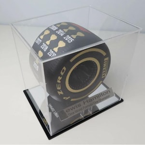 May include: A black and gold Pirelli Formula 1 tyre in a clear acrylic display case. The tyre has "ZERO" and the years 2008, 2014, 2015, 2017, 2018, 2019, and 2020. A silver plaque reads "Lewis Hamilton Time World Formula One Champion."