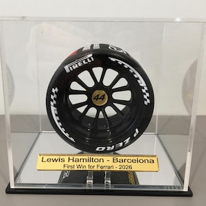 Puede incluir: Un neumático de Fórmula 1 en miniatura de color negro con la marca blanca Pirelli y P Zero, que presenta un tapacubos dorado con el número 44, dentro de una vitrina de acrílico transparente con una placa dorada Lewis Hamilton - Barcelona First Win for Ferrari - 2026.