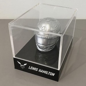 Może przedstawiać: Srebrny miniaturowy kask wyścigowy w przezroczystej akrylowej gablocie. Kask ma szczegółowe detale i logo Mercedes-Benz. Czarna podstawa gabloty ma biały napis "LEWIS HAMILTON".