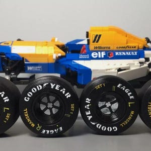 May include: A blue, white, and yellow Williams Renault Formula 1 race car model with four black Goodyear tires. The tires have white lettering that reads "Goodyear" and "Mansell". The car has the number "5" on the side.