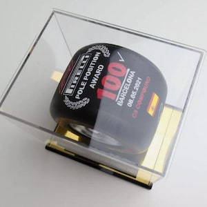 Puede incluir: Un neumático Pirelli negro con el texto "POLE POSITION AWARD" y "100 BARCELONA 08.05.2021 C3 COMPOUND" está encerrado en una caja de acrílico transparente. El neumático tiene una bandera española y se exhibe sobre un soporte dorado y negro.