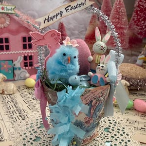 May include: An Easter-themed arrangement in a decorative cup, featuring a blue chick, pink and white bunnies, and a pink egg. The cup has a vintage illustration and is adorned with blue ribbons. A banner reads "Happy Easter!"