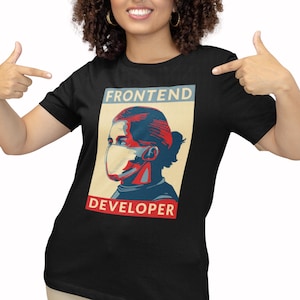 May include: Black t-shirt featuring a stylized illustration of a person wearing a face mask. The image has the words "FRONTEND DEVELOPER" in a retro poster style. The person is pointing at the shirt.
