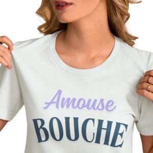 May include: A light-colored t-shirt with the words "Amouse Bouche" printed on the front in a two-tone design. The word "Amouse" is in a light purple script, and "Bouche" is in a dark blue sans-serif font.