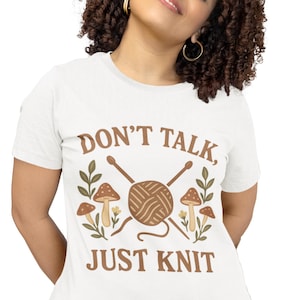 May include: White t-shirt featuring the text "DON'T TALK, JUST KNIT" in brown. The design includes a ball of yarn, knitting needles, mushrooms, and foliage. A casual tee for knitters.