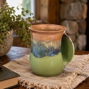 May include: A ceramic mug featuring a multi-toned glaze with a light green base, a dark blue and cream middle section, and a tan rim. The mug has a wide, flat, integrated handle extending from the side, creating a smooth, continuous silhouette.