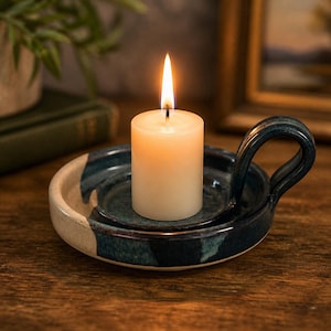 May include: A lit, off-white candle sits in a handmade, ceramic candle holder. The candle holder is a dark teal color with a handle and a cream-colored rim. The candle flame is bright yellow.