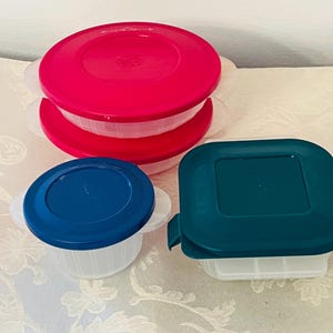 May include: A collection of plastic food storage containers with colorful lids. The containers are white with red, blue, and teal lids. Some have handles, and one is square-shaped.